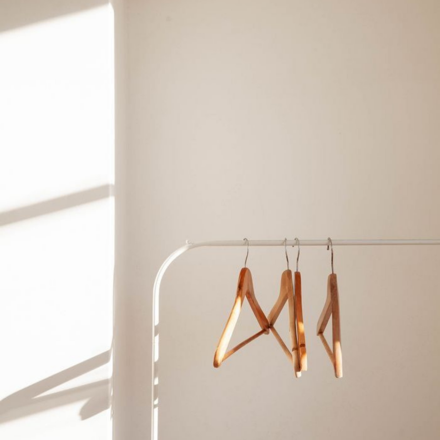 A few empty wooden hangers on a white rail against a cream wall, in soft window light.