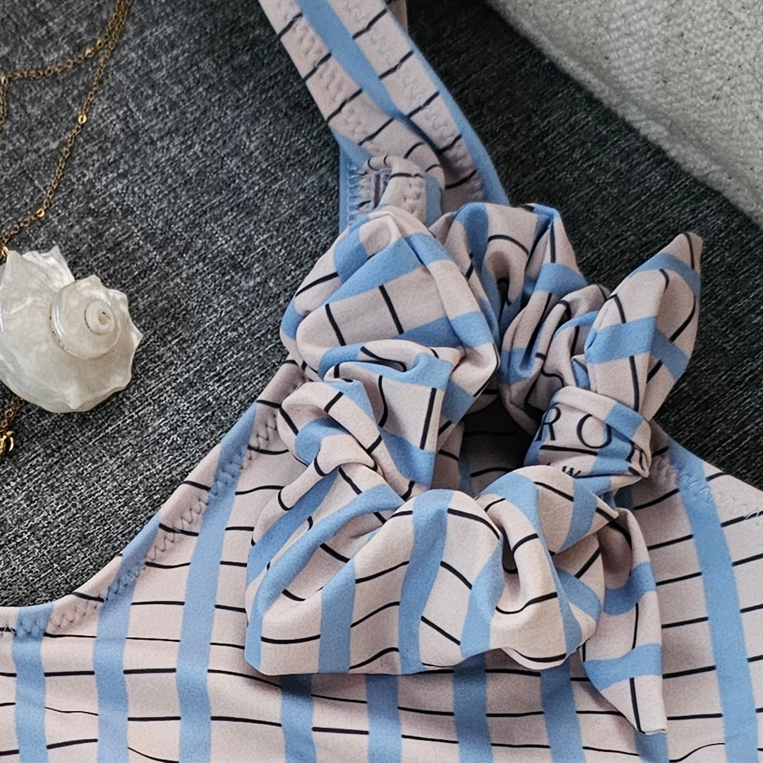 A Laguna check scrunchie in sand and soft blue resting on the matching bikini, beside a shell pendant.