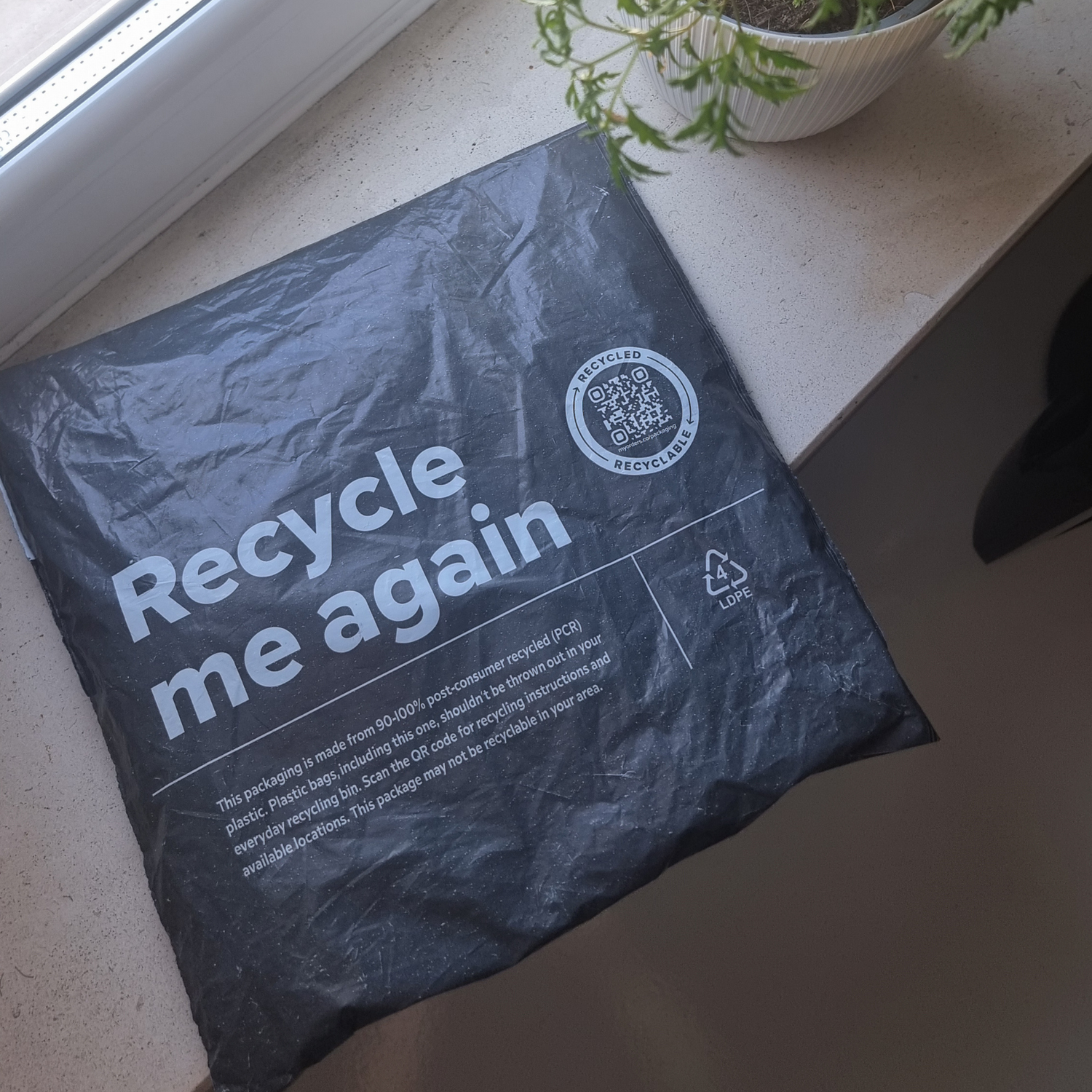 A grey mailer printed "Recycle me again", made from 90–100% post-consumer recycled plastic, on a windowsill beside a potted plant.