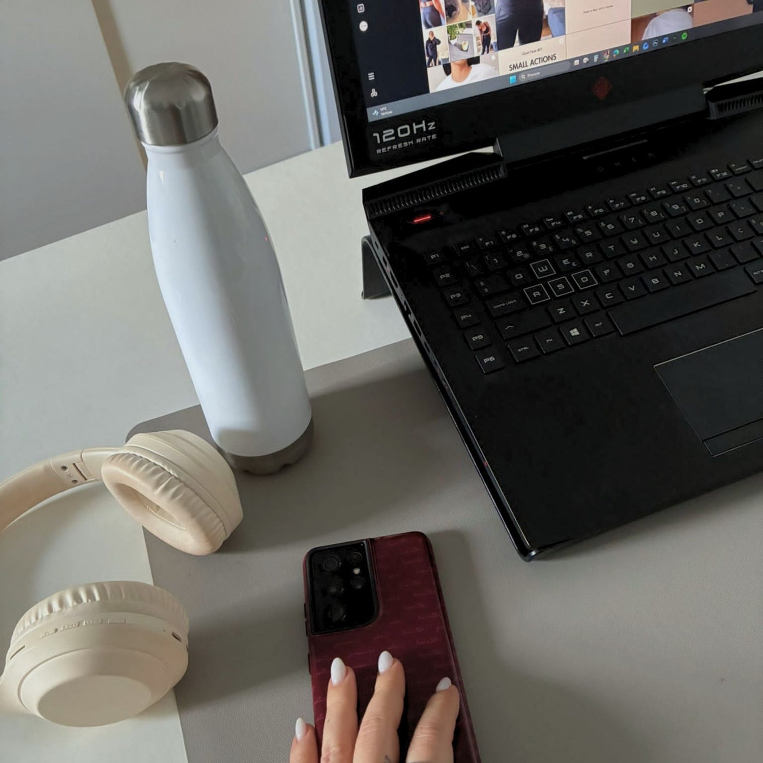 A desk with a laptop, a white water bottle, headphones and a phone, one hand resting on the screen — where Aurora is made, day to day.