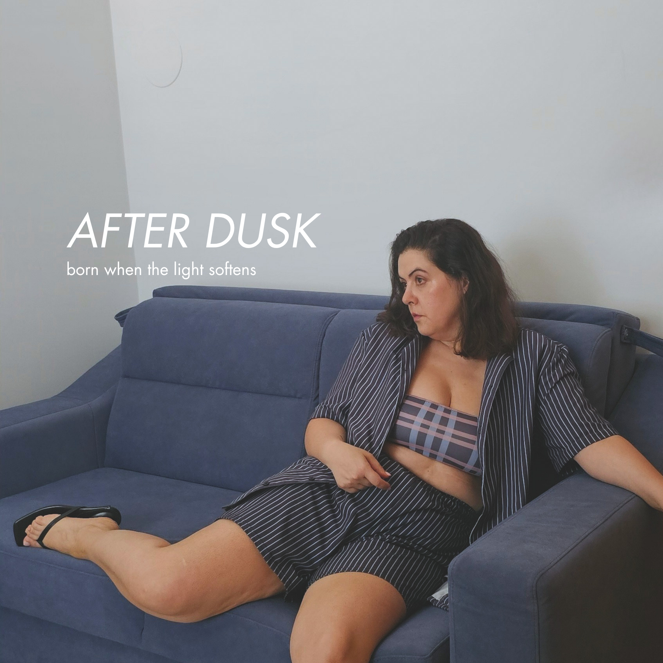 Woman relaxing on a navy blue sofa, wearing an open pinstripe shirt and matching shorts over a plaid bikini top, for the Aurora Soulwear After Dusk collection, born when the light softens.
